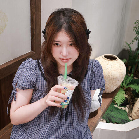 woman with long hair drinking from a plastic cup. She looks down and out of frame, there is a large vase behind her